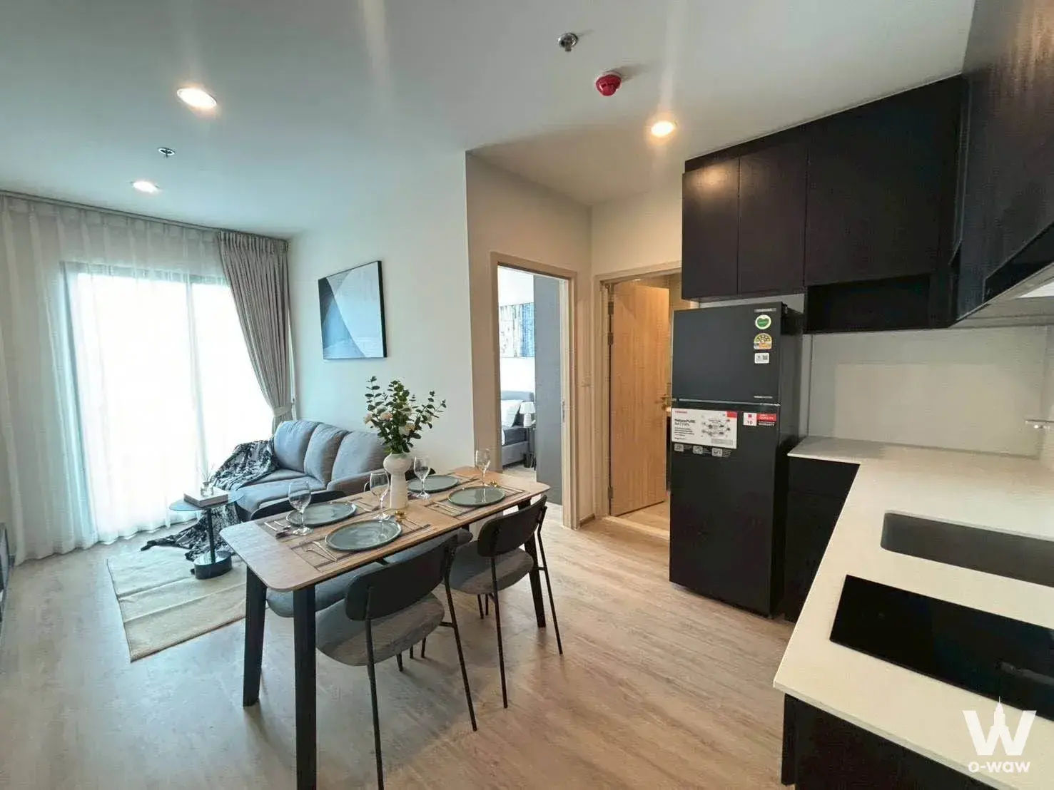Open-plan living and dining area in a Nue District R9 condo with grey seating, wood-grain table, and black kitchen accents
