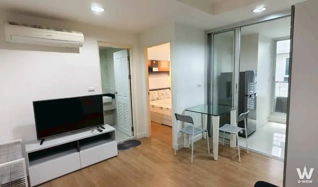 Modern open-plan living room in The Kris 5 Ratchada 17 condo with light wood floors and a glass kitchen divider
