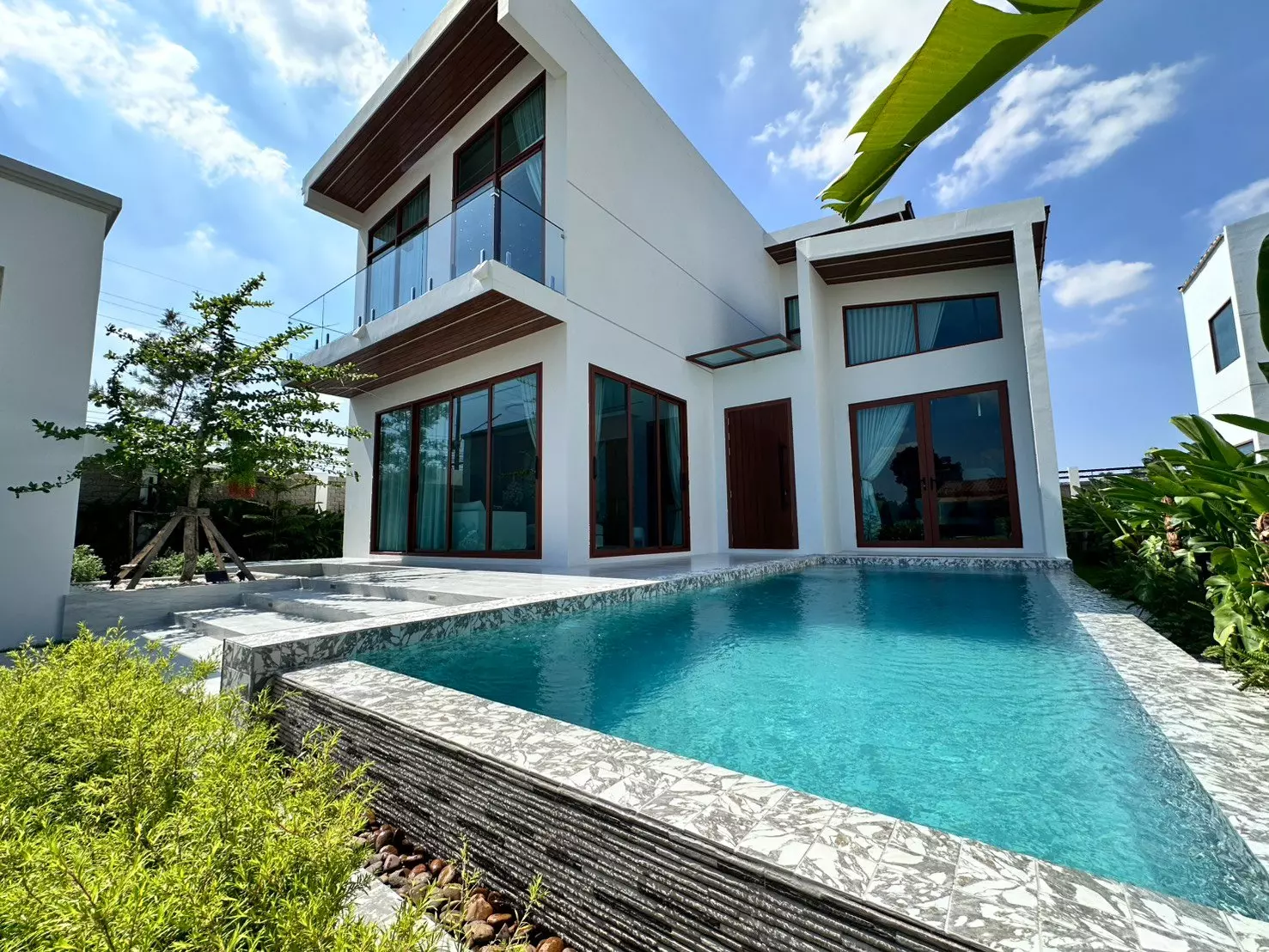 Exterior of modern white villa for sale in Pattaya with turquoise pool, dark wood accents, and tropical garden