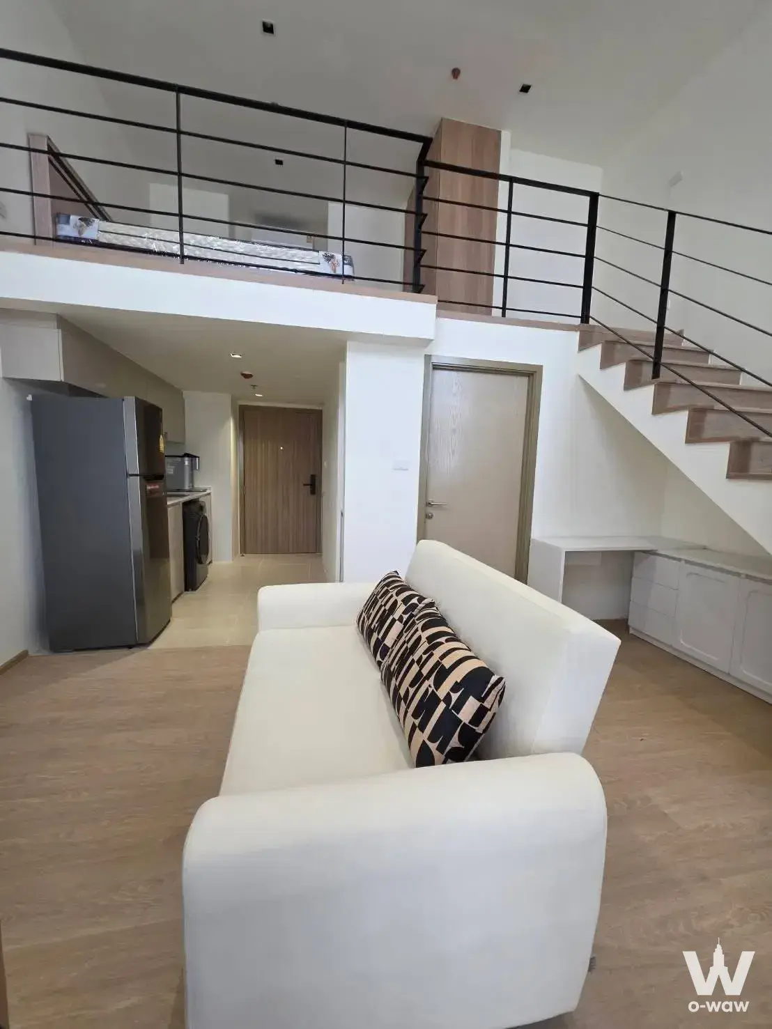 Open-plan duplex living area with white sofa, kitchenette, and floating staircase leading to mezzanine