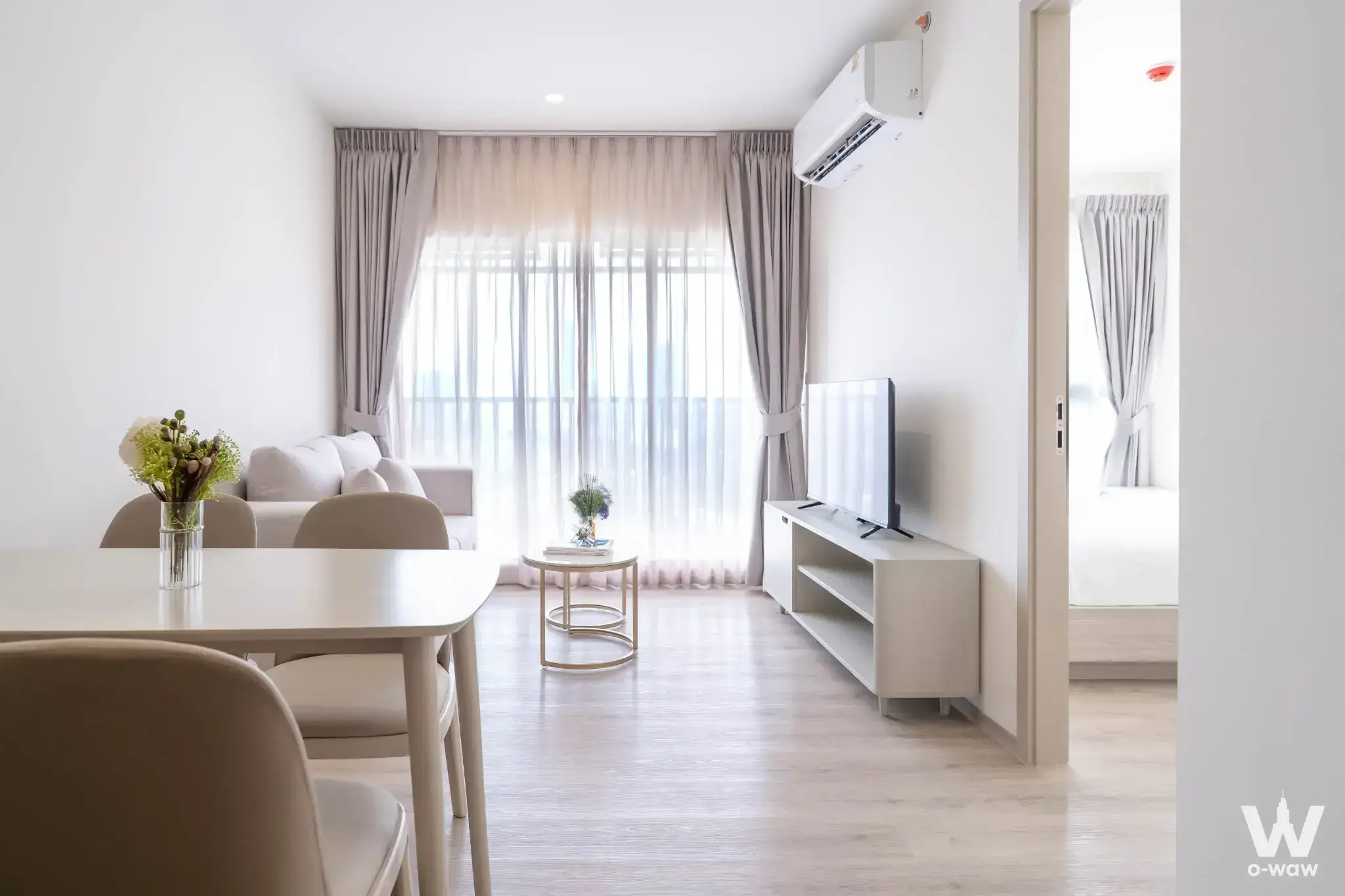 Open-plan living area in this Origin Place Bangna condo features light wood floors and a large balcony with sheer curtains