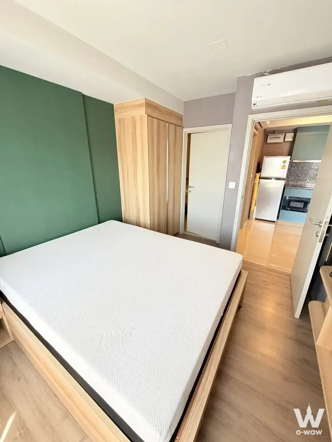 Studio bedroom in THE BASE Sukhumvit 50 showing a queen bed, oak wardrobe, and view into the kitchen