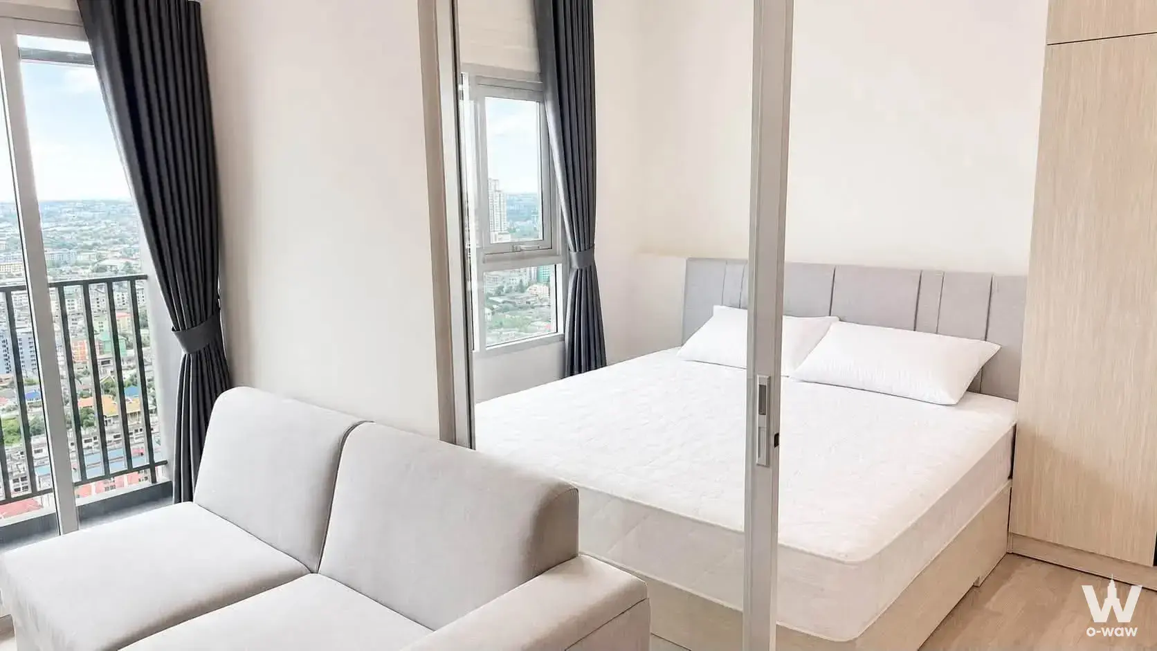 Studio at Niche MONO Ramkhamhaeng with beige sofa, bed behind glass partition, and floor-to-ceiling window