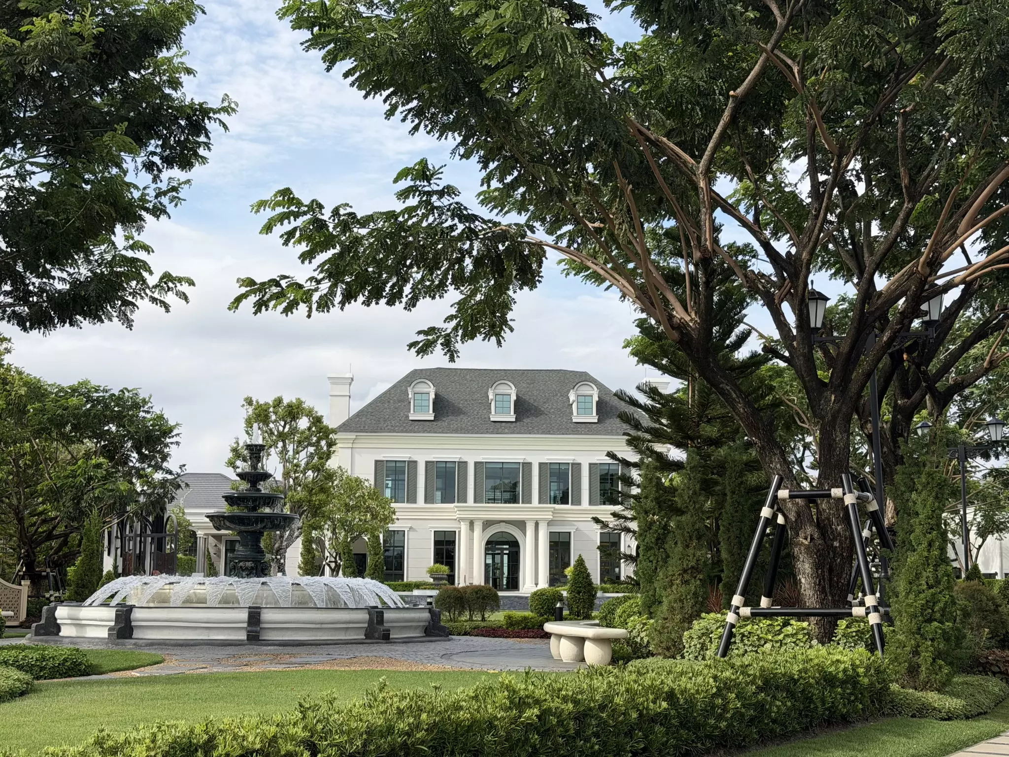 White mansion exterior with grey mansard roof, dormer windows, and ornate fountain in Setthasiri Bangna