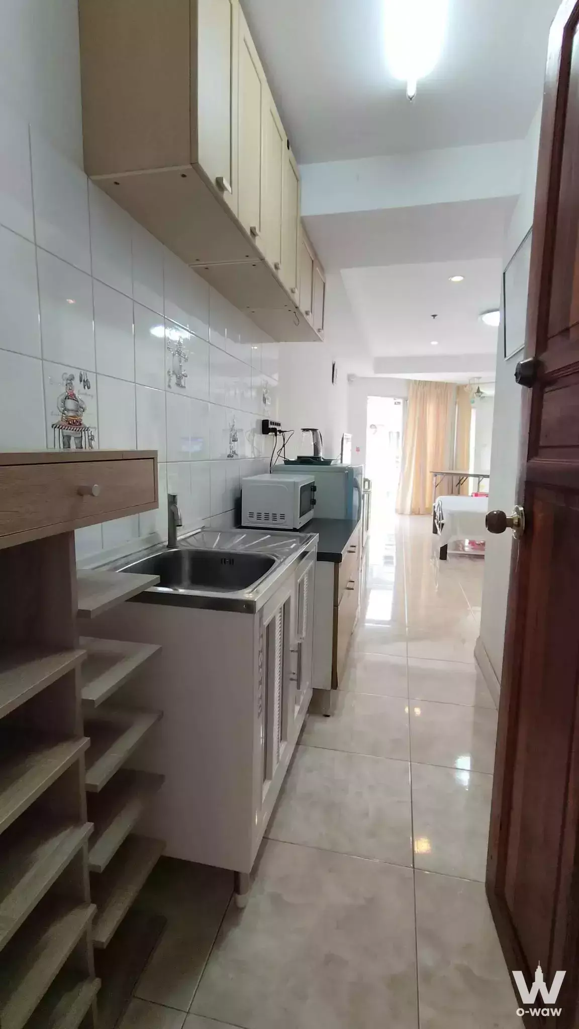 Galley kitchen in 1-bed Rimhad Condo with wood cabinets, chef-motif backsplash, and view into bedroom with peach curtains