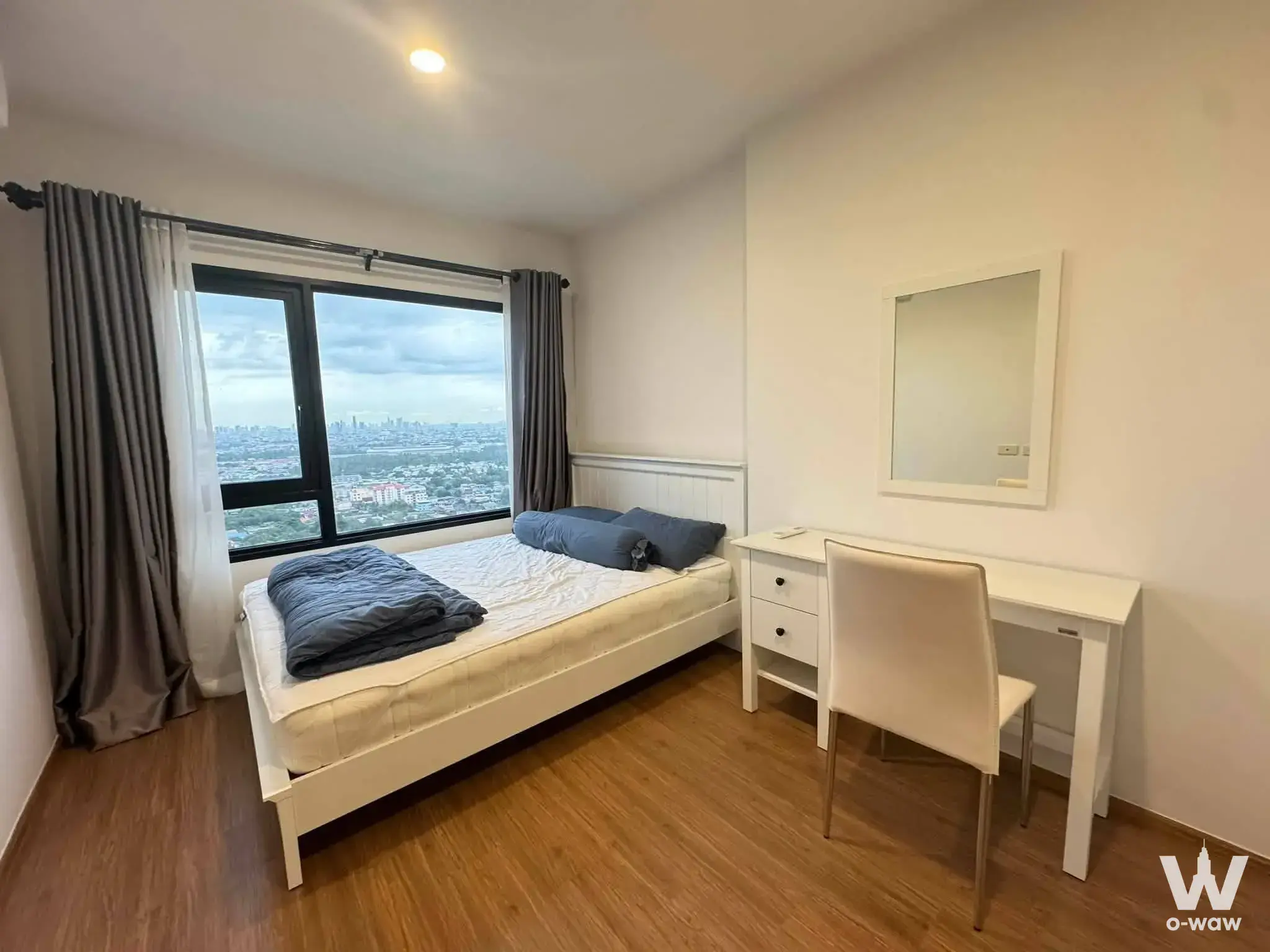 Furnished bedroom with blue bedding, desk, and city skyline view in a 1-bed condo for rent at The LIVIN Phetkasem
