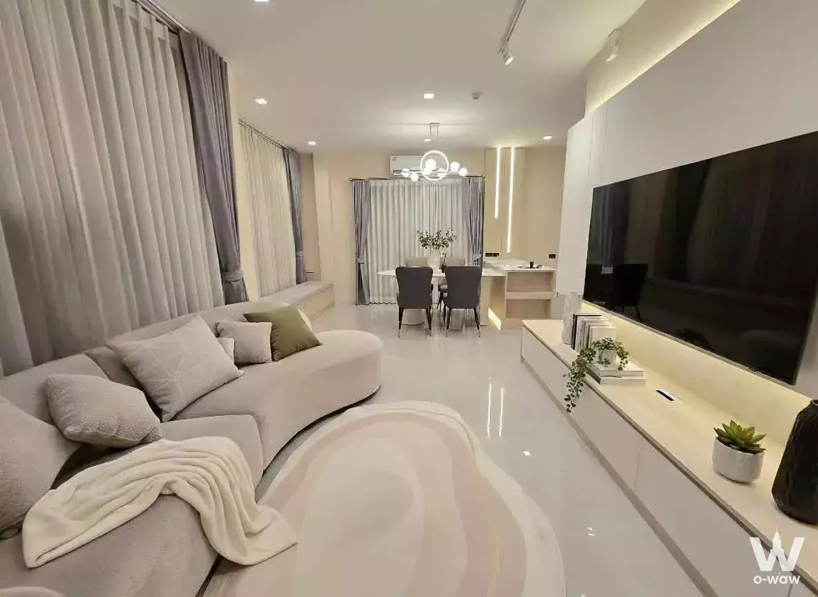 Open-plan living area in Bangkok Boulevard Bangna with curved beige sofa, wall-mounted TV, and round dining set