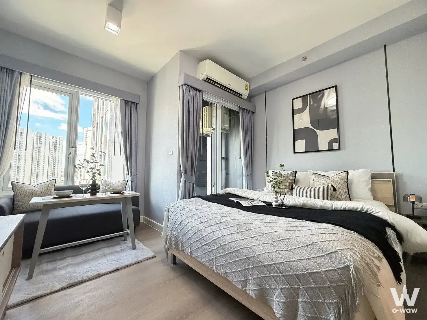 Bright bedroom with queen bed, city skyline view, light wood floors, and dark grey sofa near window