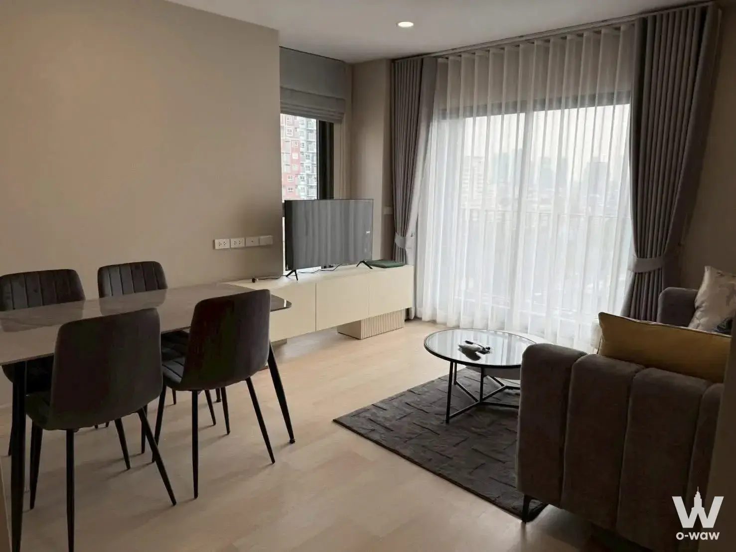 Open-plan living room in Aspire Vibha-Victory condo featuring grey sofa, dining set, and high-rise city views