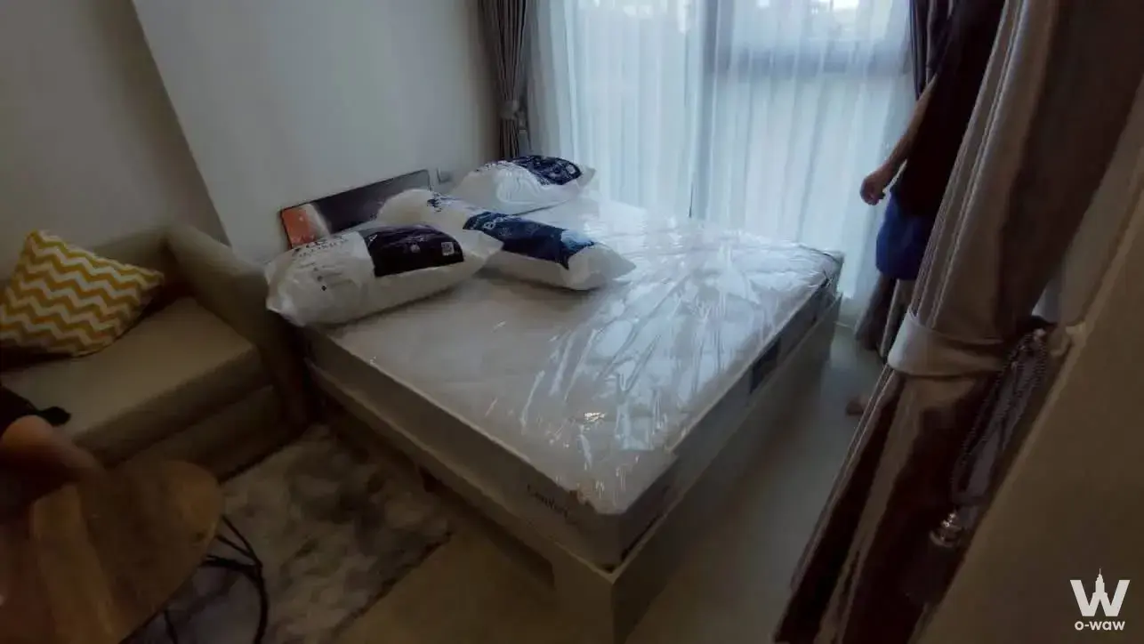 Compact bedroom in Atmoz Oasis Onnut showing new bed frame, beige loveseat, yellow pillow, and large window with drapes