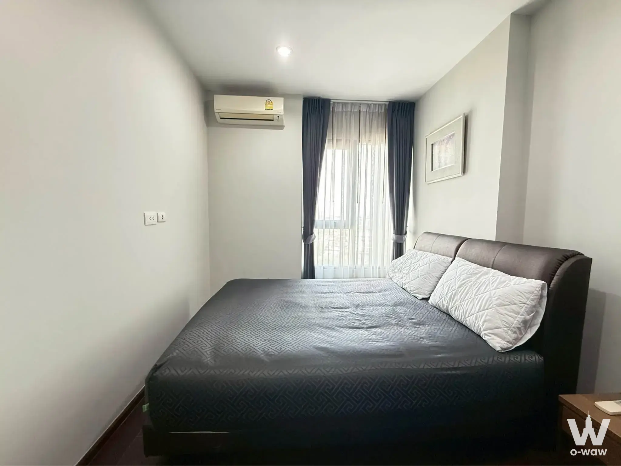 Bedroom in 1-bed C Ekkamai Condominium with dark leather bed frame, grey duvet, white walls, and blue drapes