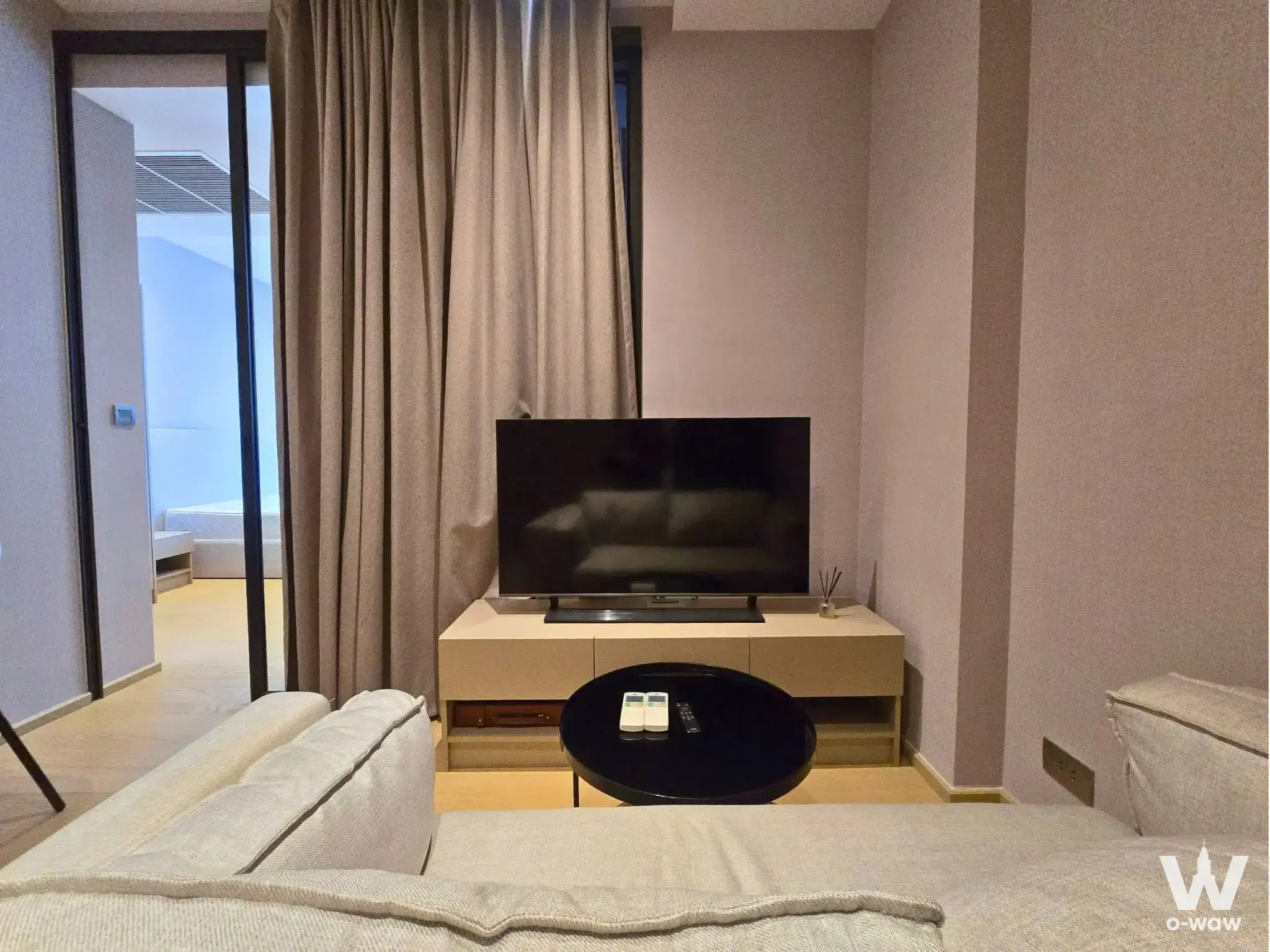 Living room in a 1-bed ASHTON Asoke condo with beige sofa, media console, and open bedroom door