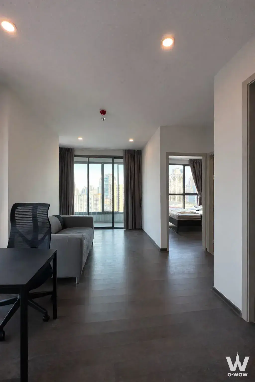 Living room in the Ideo Q Siam-Ratchathewi condo features a grey sofa and sliding glass doors with a city skyline view