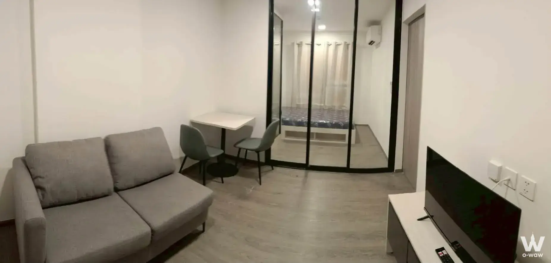 Bright studio with grey sofa, sage chairs, and glass partition separating bedroom in Kave Wonderland condo