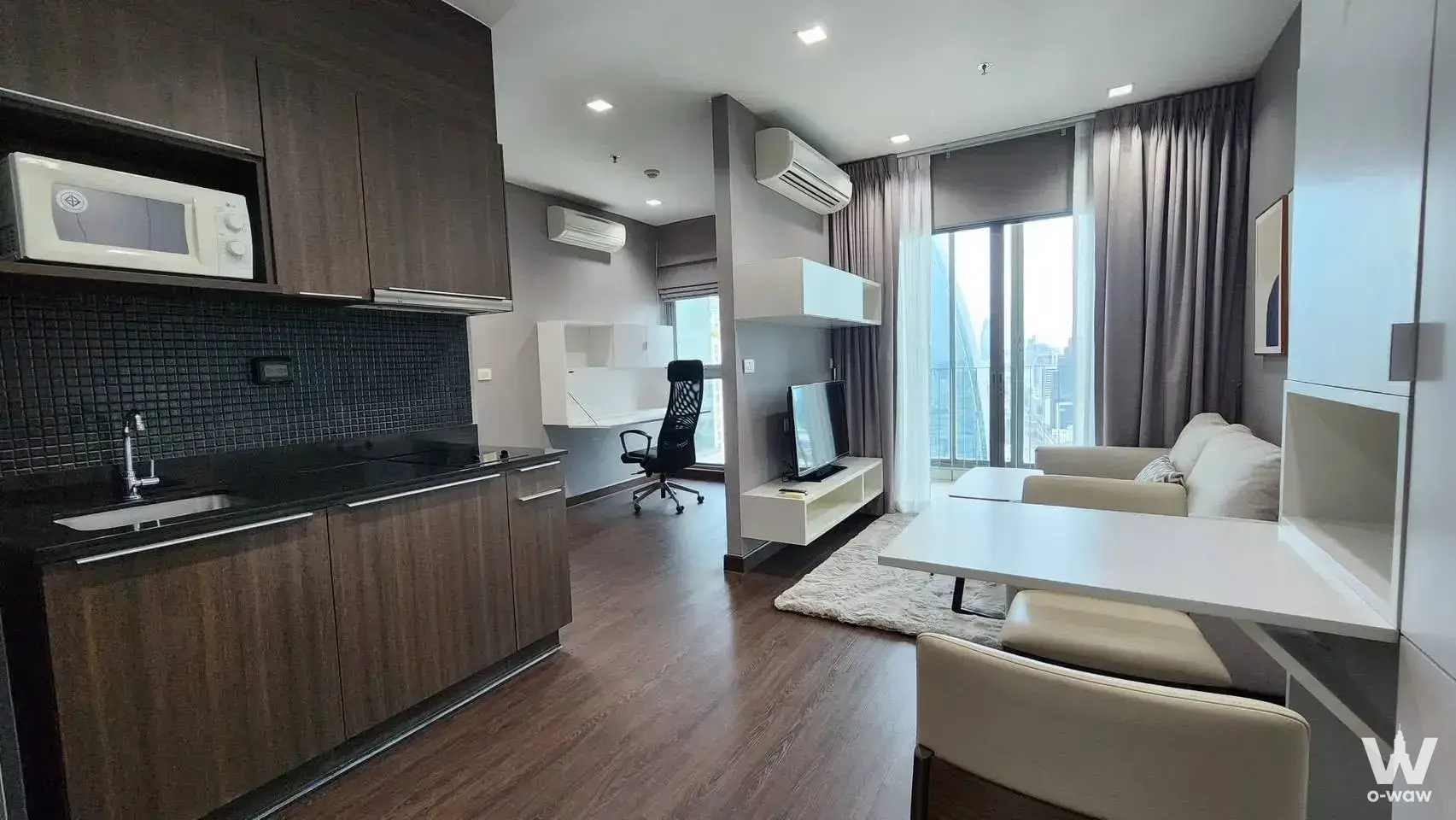 Open-plan studio with dark wood kitchenette, grey partition wall, beige sofa, and high-rise city view windows