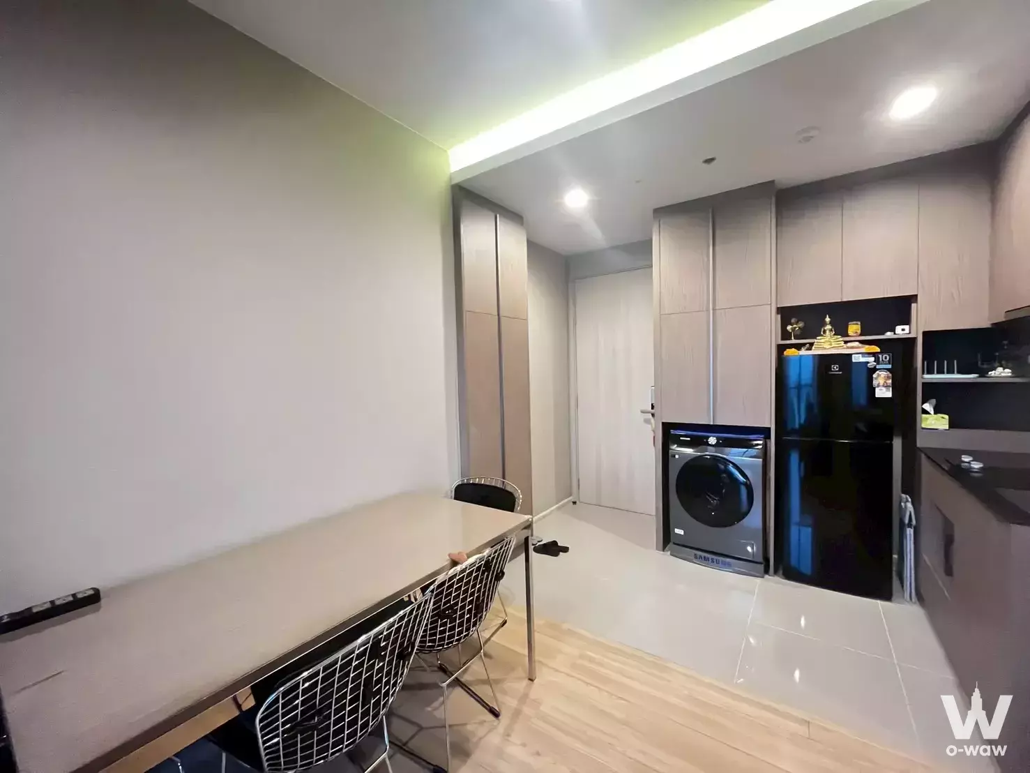 Open-plan dining area in M Jatujak condo featuring a light wood table and integrated kitchen appliances
