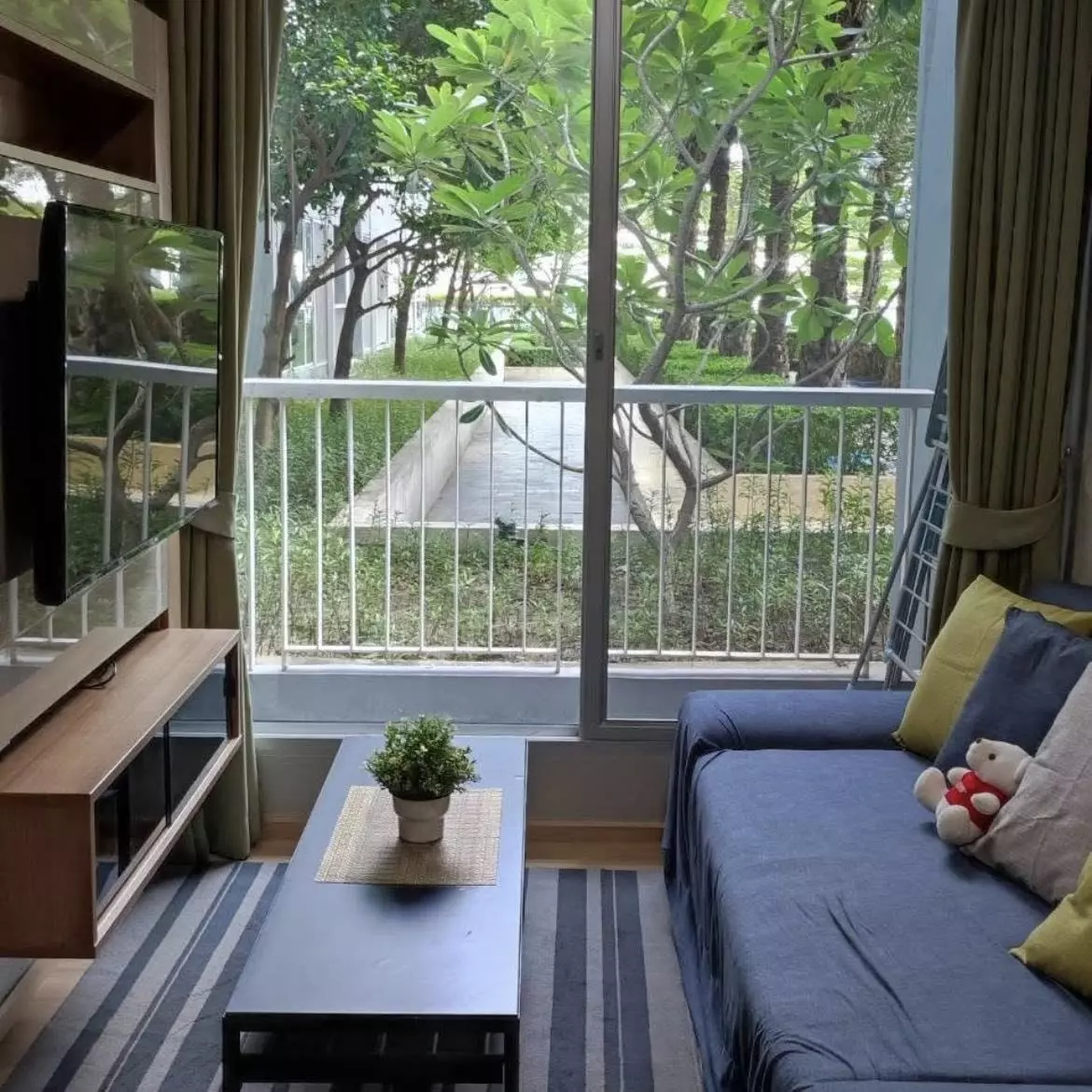 Living room with dark blue sofa and lush green tree view in a 1-bed condo for rent in Rhythm Sukhumvit 50