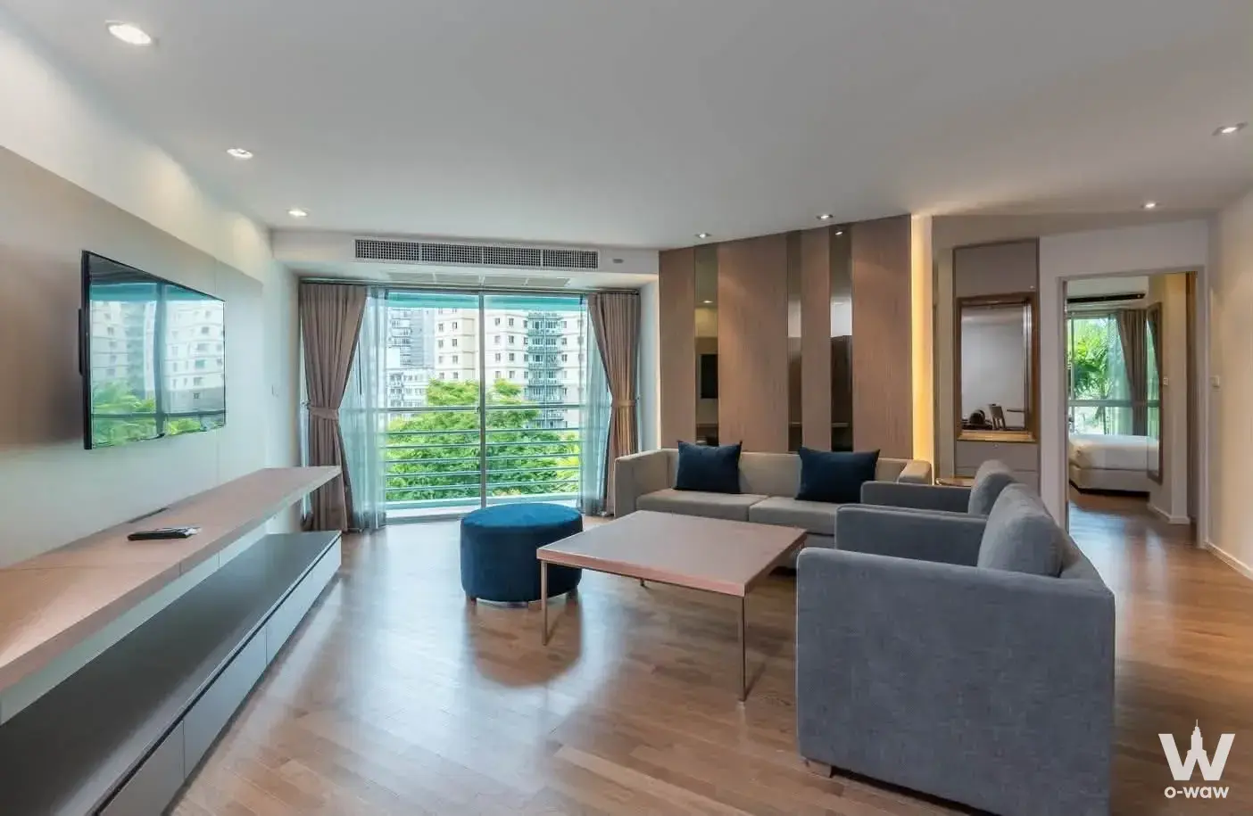 Spacious living room with grey sofa, wall-mounted TV, and balcony access offering city views in a Bangkok Garden Apartment