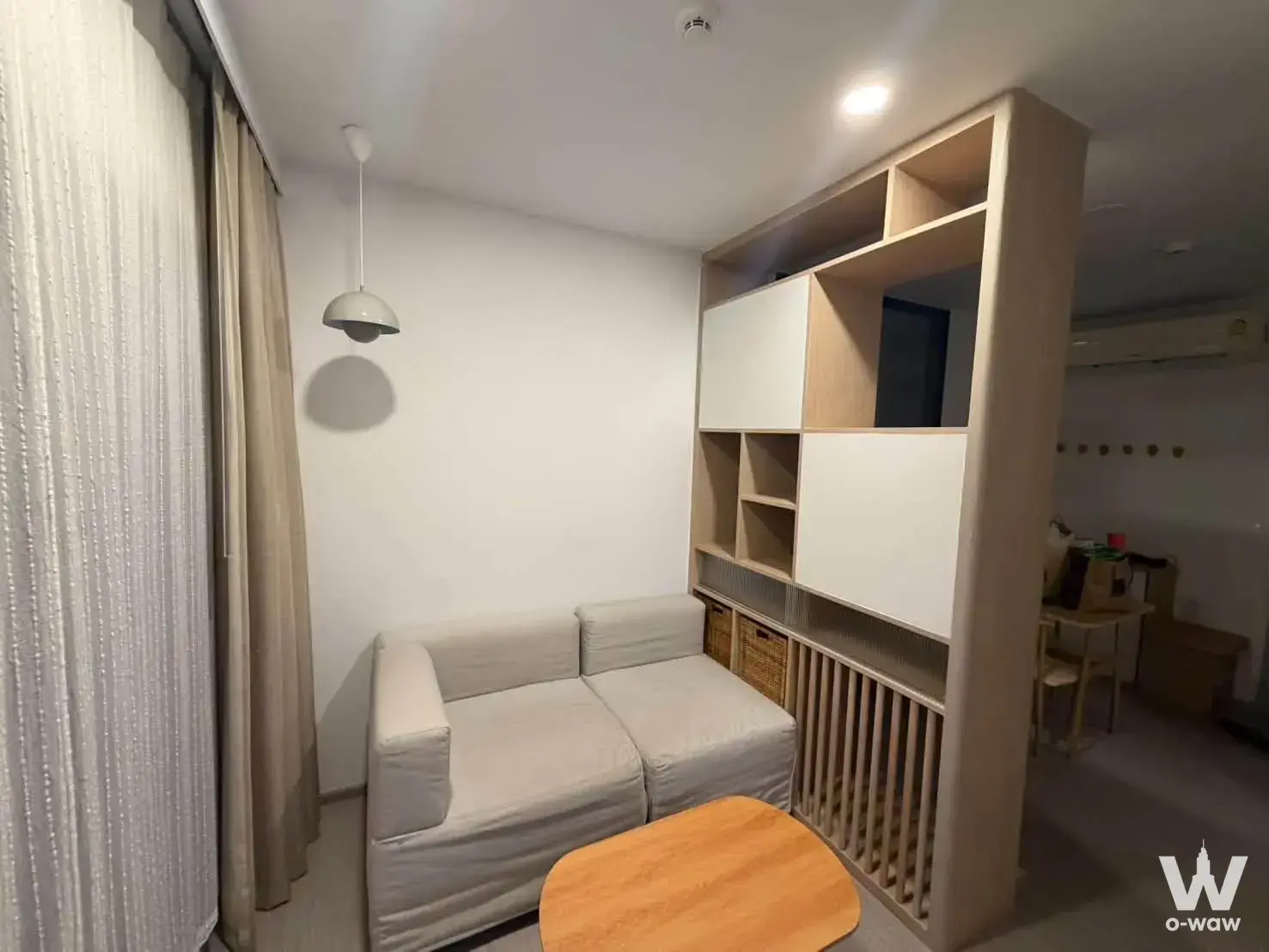 Minimalist studio living area with fabric sofa, open shelving divider, and pendant lighting in a 1-bed Aspire Sukhumvit-On