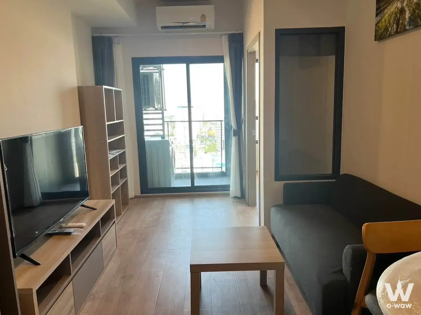 Living room in a 1-bed IDEO Rama 9 condo with a dark grey sofa, light wood media console, and balcony access
