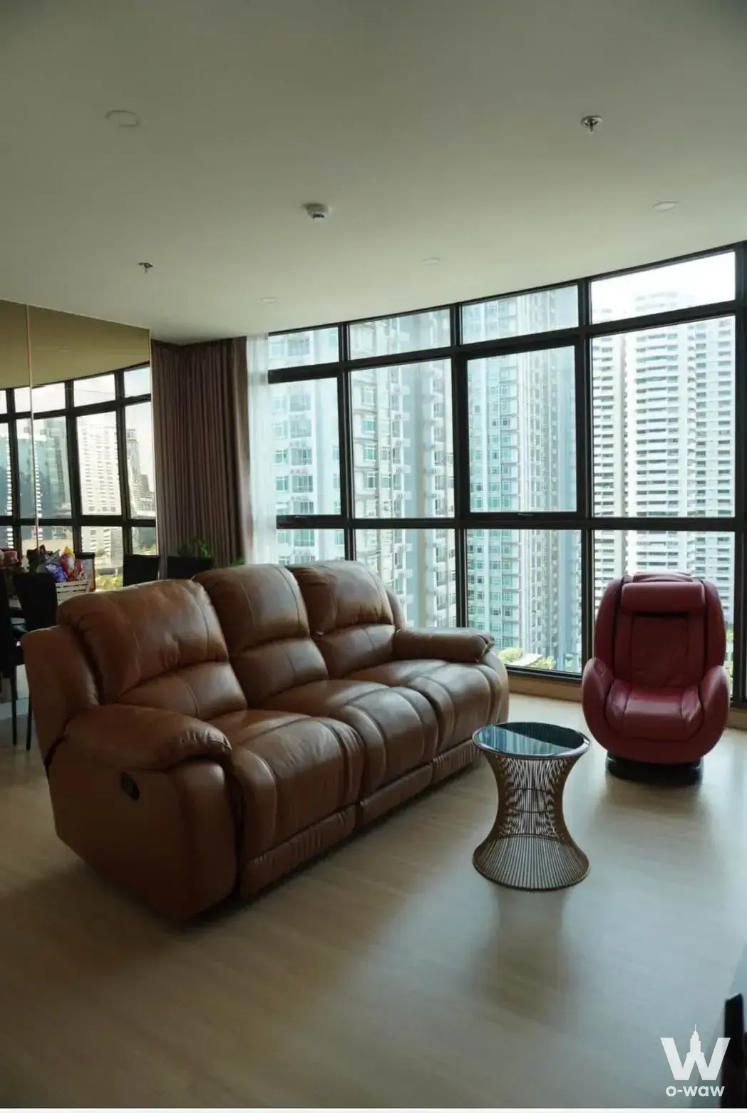 Makkasan condo living area featuring a brown leather sofa and red massage chair by floor-to-ceiling windows