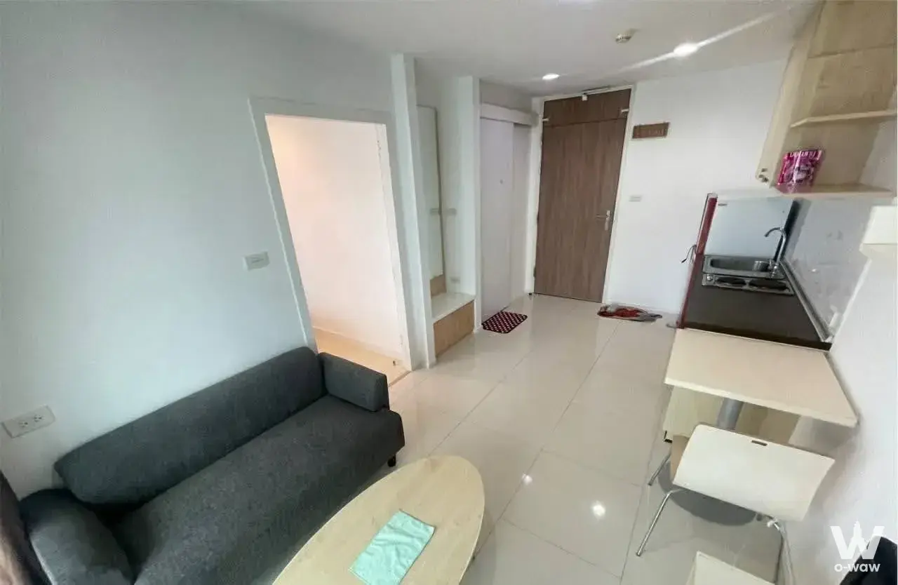 Studio living area in a Punnawithi condo with grey sofa, compact kitchenette, and glossy white tile floor