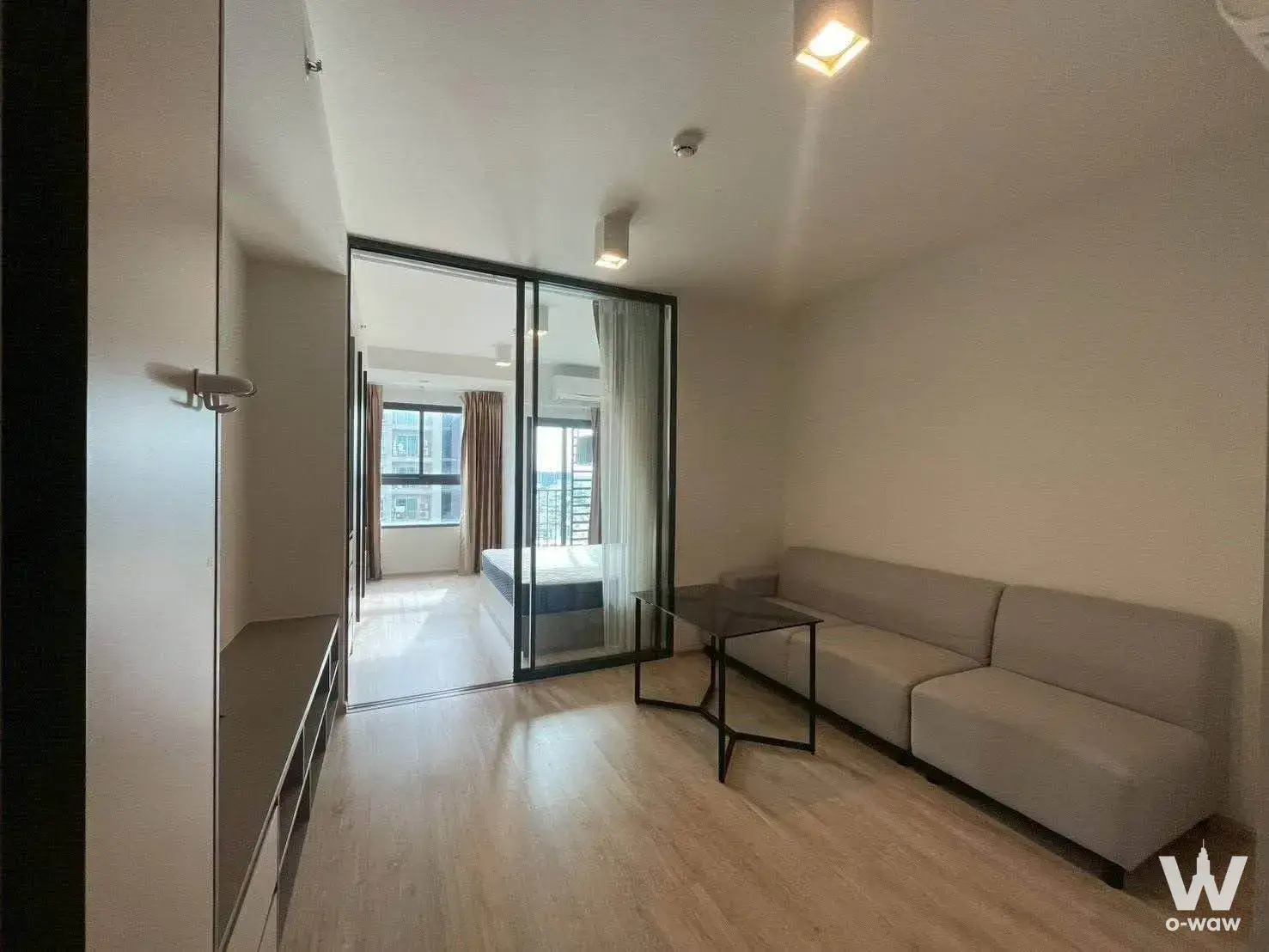 Living area with a beige sofa and glass partition revealing the bedroom in Ideo Sathon-Wongwian Yai