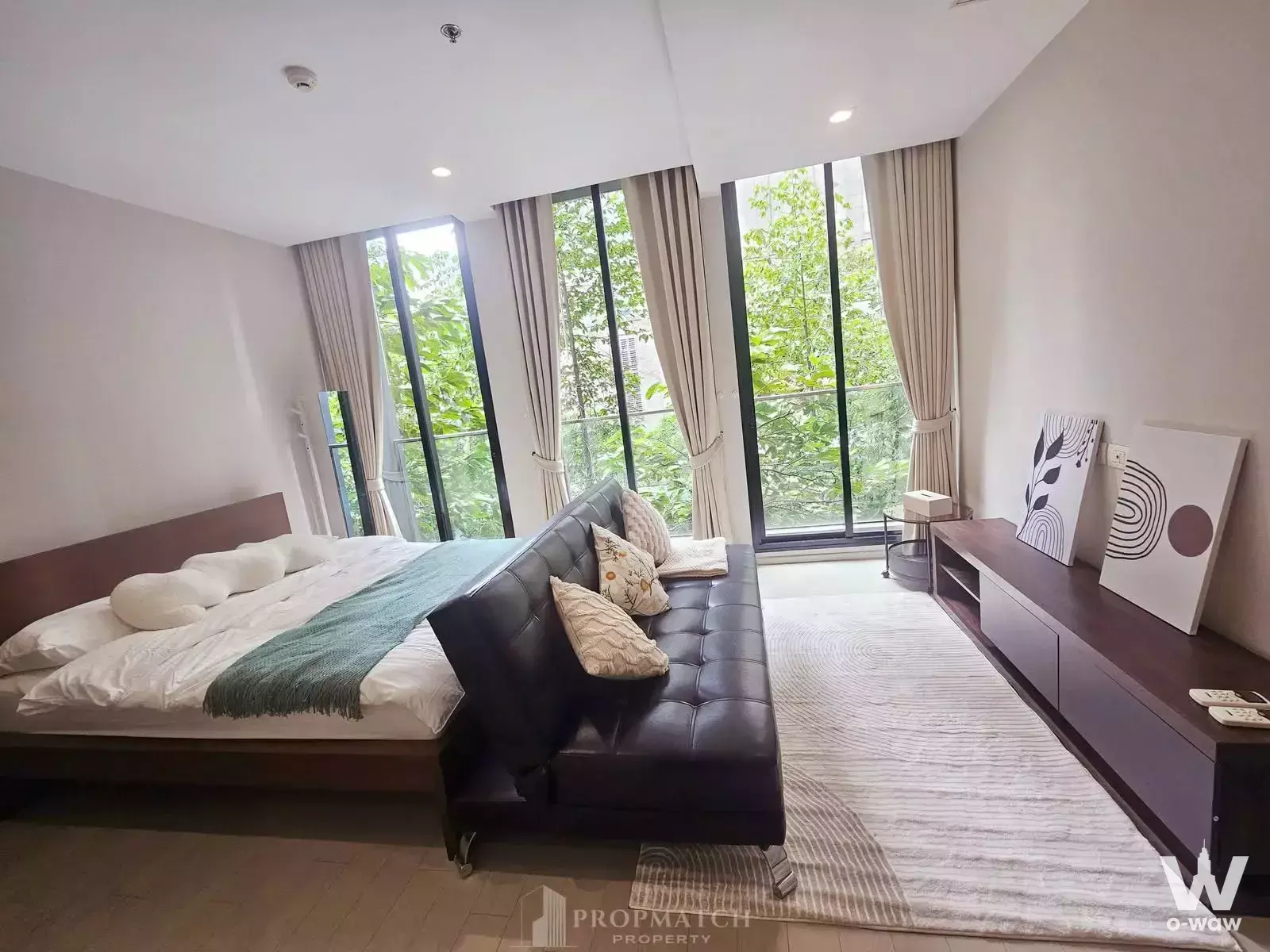 Bedroom in Noble Ploenchit condo with a queen bed, dark wood accents, and floor-to-ceiling windows overlooking greenery