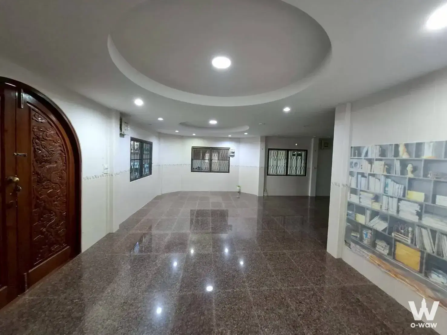 Empty lobby with polished granite floors, arched wood door, and floor-to-ceiling bookshelf in Bang Na Villa Village