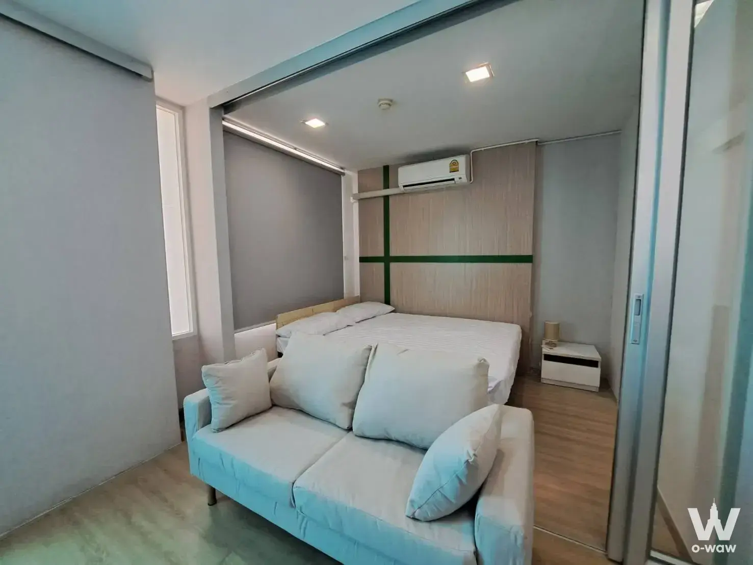 Open-plan studio in The Colory Vivid with grey sofa and wood-paneled bedroom wall