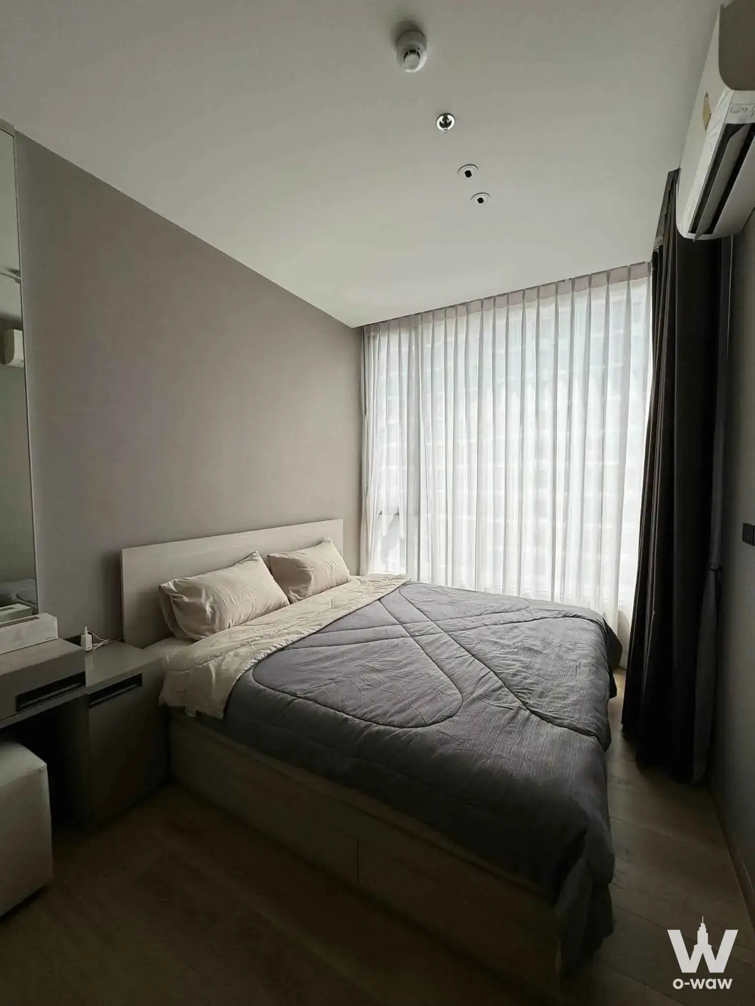 Minimalist bedroom in The Fine Bangkok condo featuring light wood floors, grey bedding, and built-in desk