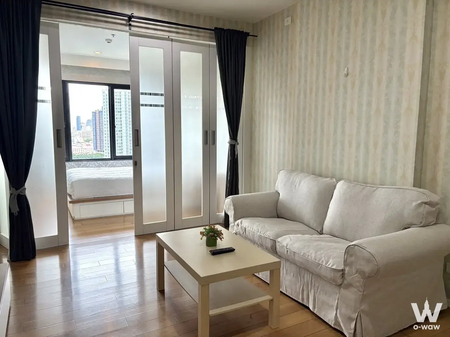 Living room in a Blocs 77 1-bed condo featuring beige sofa and frosted glass partition to bedroom
