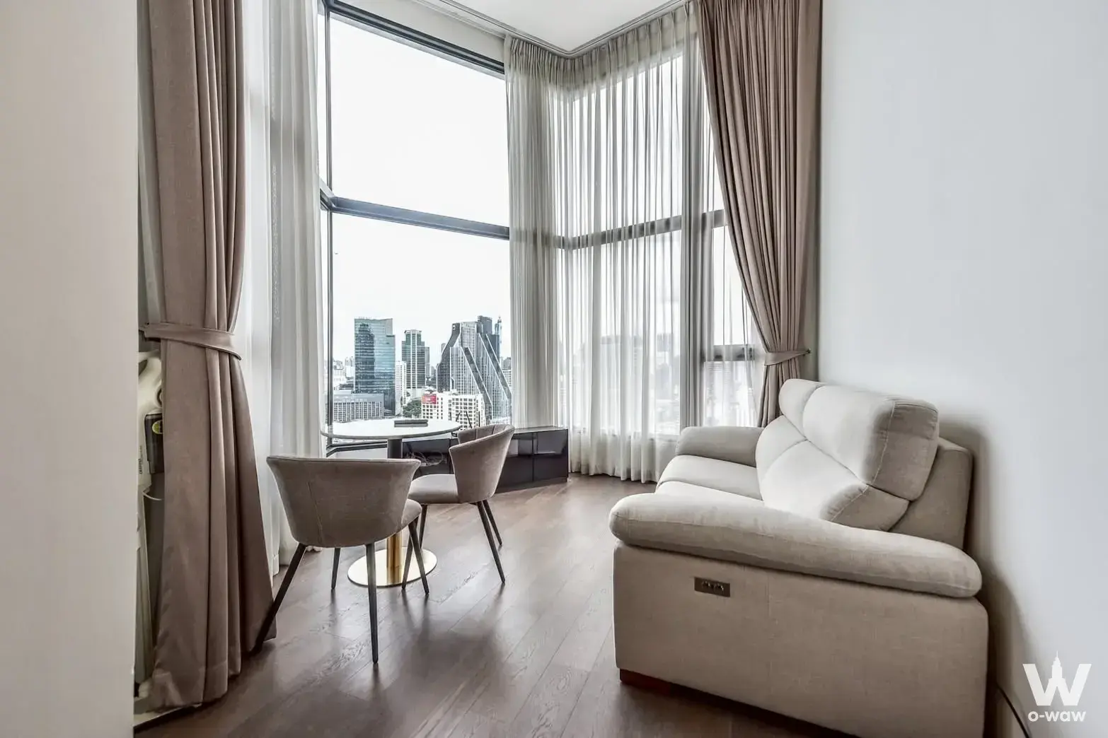 Cream recliner sofa and taupe chairs face floor-to-ceiling windows with a panoramic city view in this 2-bed Park Origin
