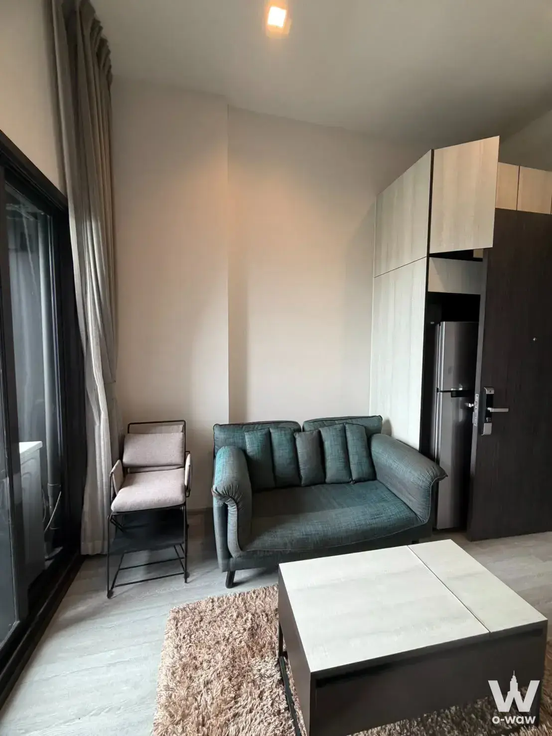Furnished living room in THE LINE Sukhumvit 71 with teal sofa, floor-to-ceiling windows, and built-in fridge