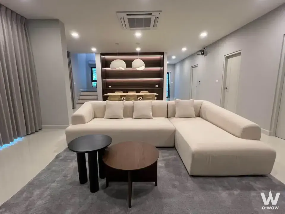 Living room in this Centro Bangna 4-bed house features a cream sectional sofa and dark wood built-in shelving