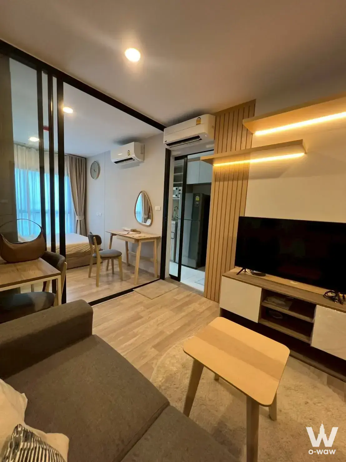 Plum Condo Sukhumvit 97.1 studio featuring grey sectional sofa, wood slat wall, and glass bedroom partition