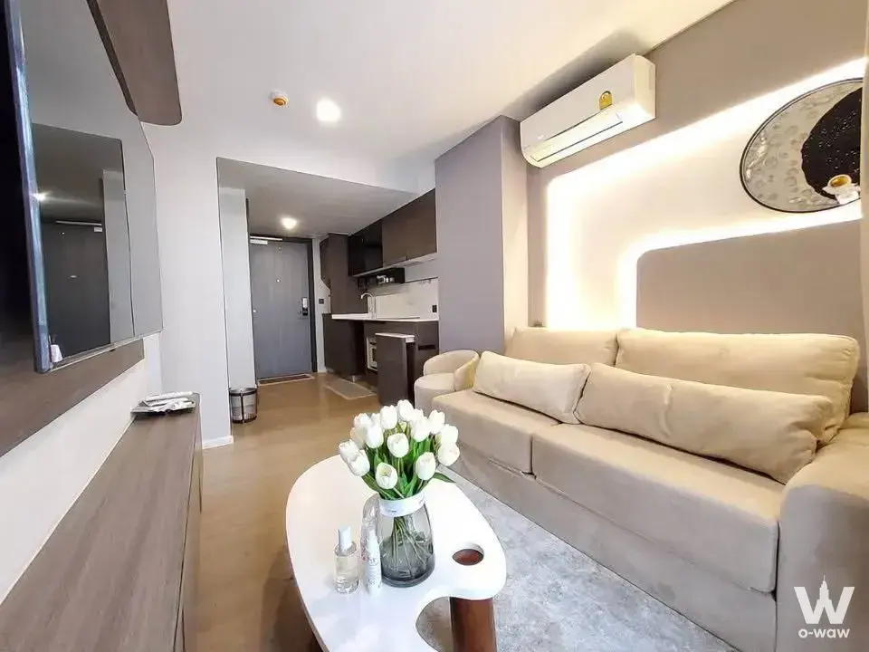 Open-plan living area in a Suanbua Residence 1-bed condo with beige sofa, taupe accent wall, and visible kitchenette