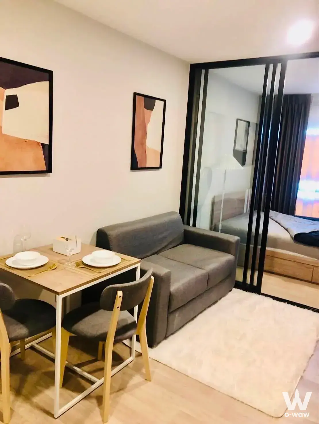 Compact studio in Plum Condo Sukhumvit 97.1 showing grey sofa, dining nook, and glass partition to sleeping area