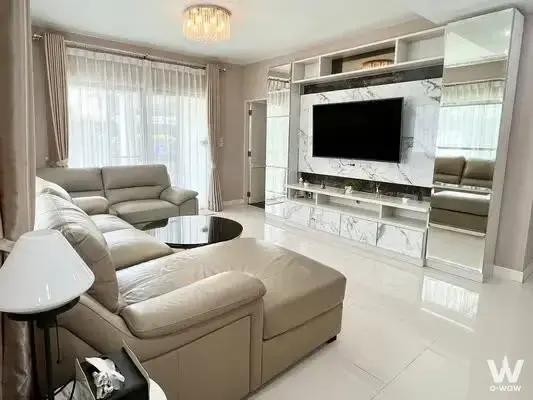 Spacious living room in a Mantana Bangna house with beige leather sectional, crystal chandelier, and floor-to-ceiling windows