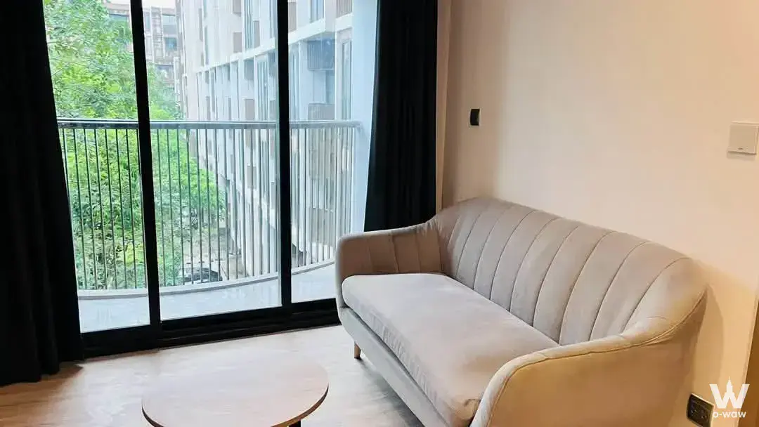 Living room in a 1-bed condo for rent in Kawa Haus has a beige velvet sofa and a balcony view of green trees