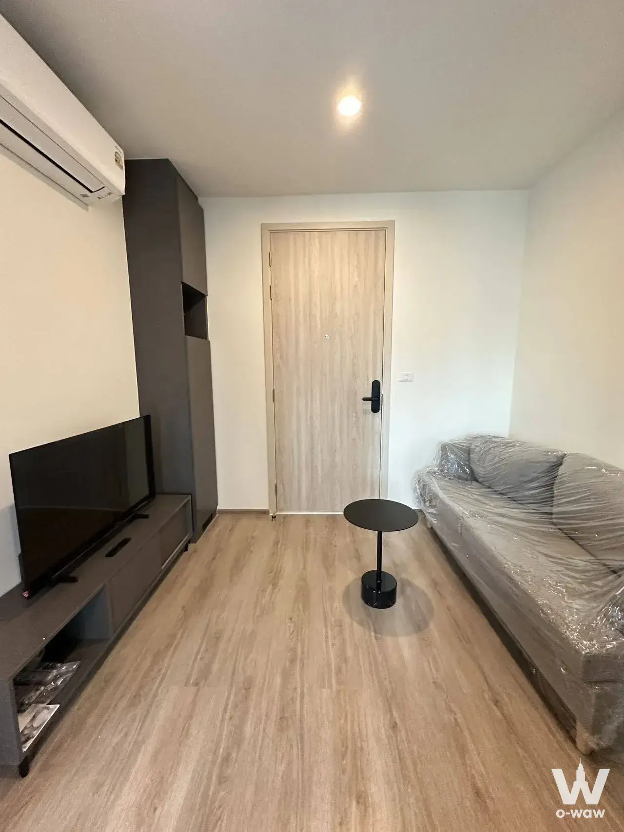 Living room in a 1-bed Tara Ruan Ake condo with oak floors, dark grey storage, and a new sofa wrapped in plastic