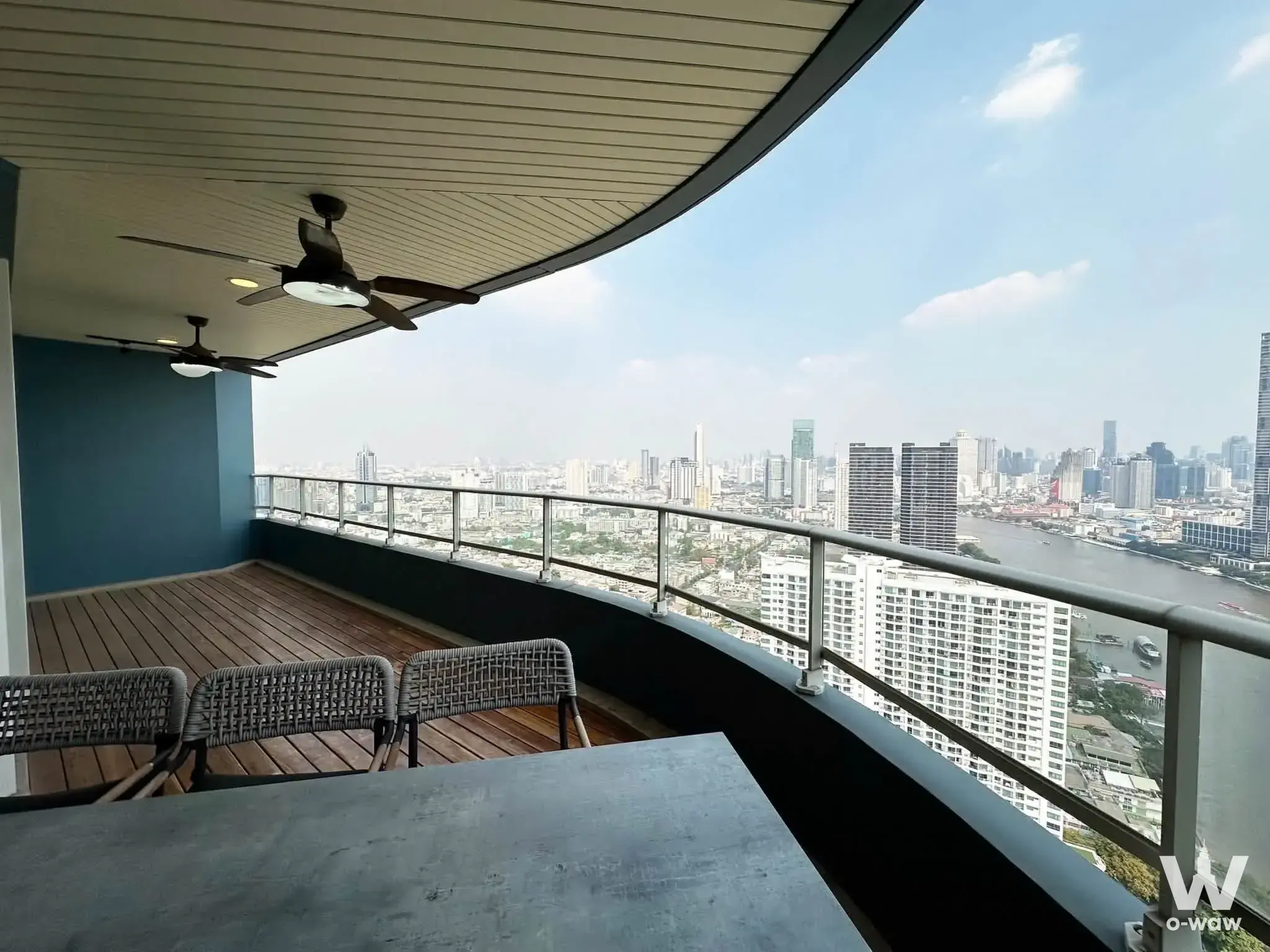 Curved balcony of a Chao Phraya condo with wood decking, woven chairs, and an expansive river skyline view