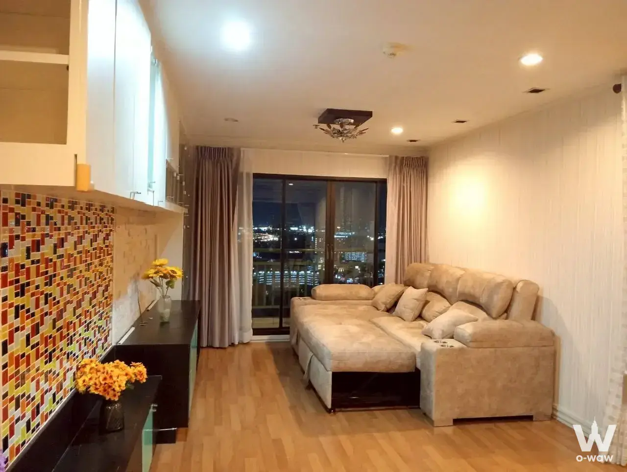 Living room in a Lumpini Place Phahon-Saphan Khwai condo with beige sectional sofa, mosaic backsplash, and night city