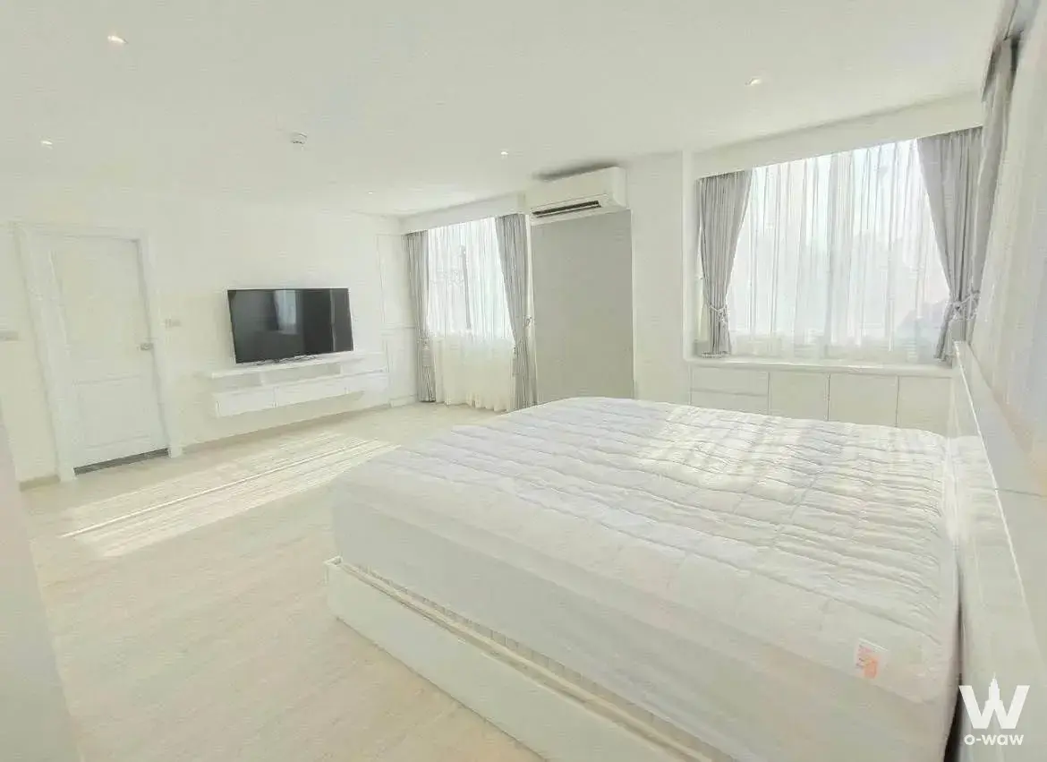 Bright master bedroom in a Silom condo featuring a white platform bed, wall-mounted TV, and large windows with sheer curtains