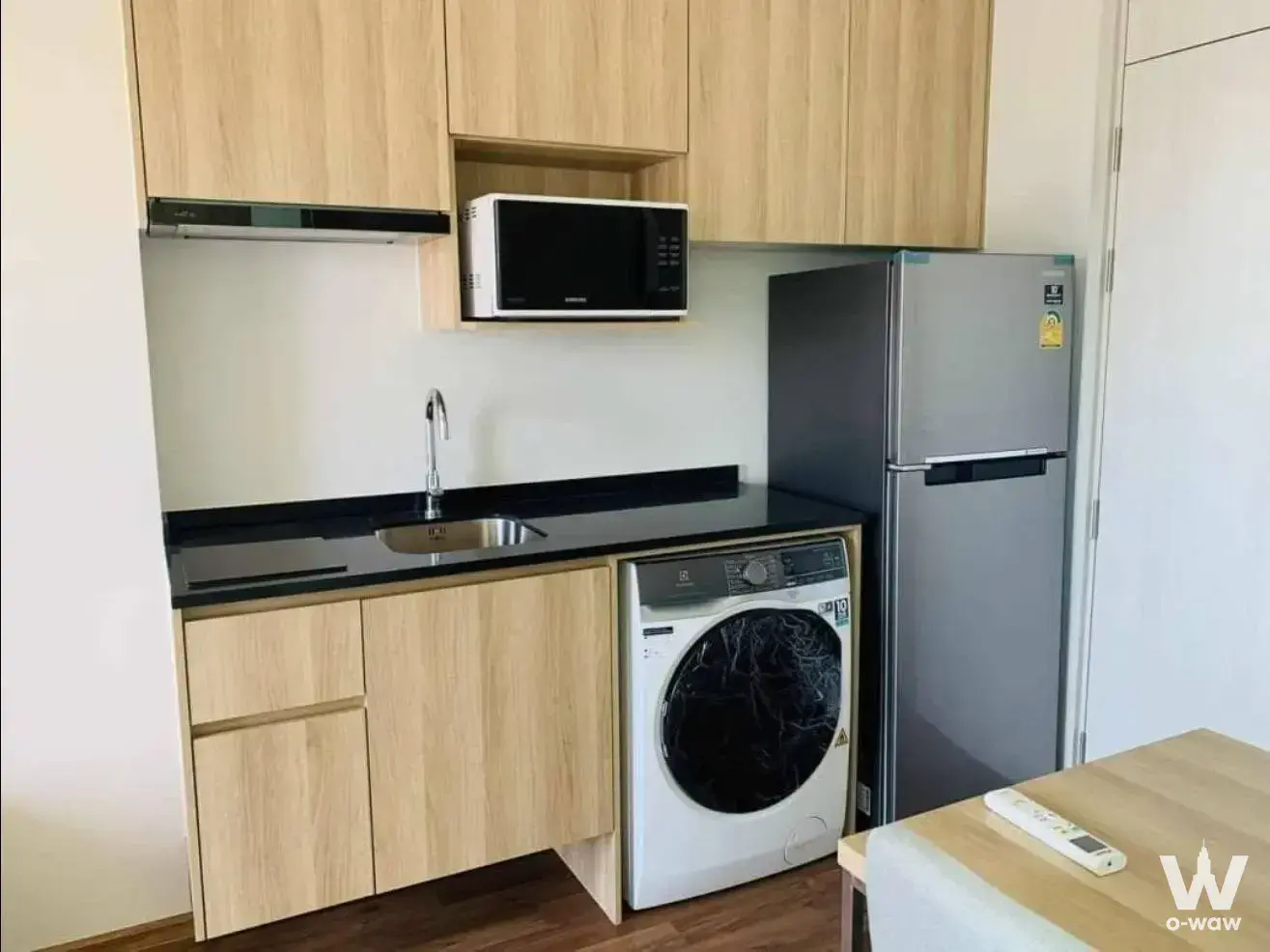 Compact kitchenette with light oak cabinets, black counter, and built-in appliances in a 1-bed condo for rent at Noble