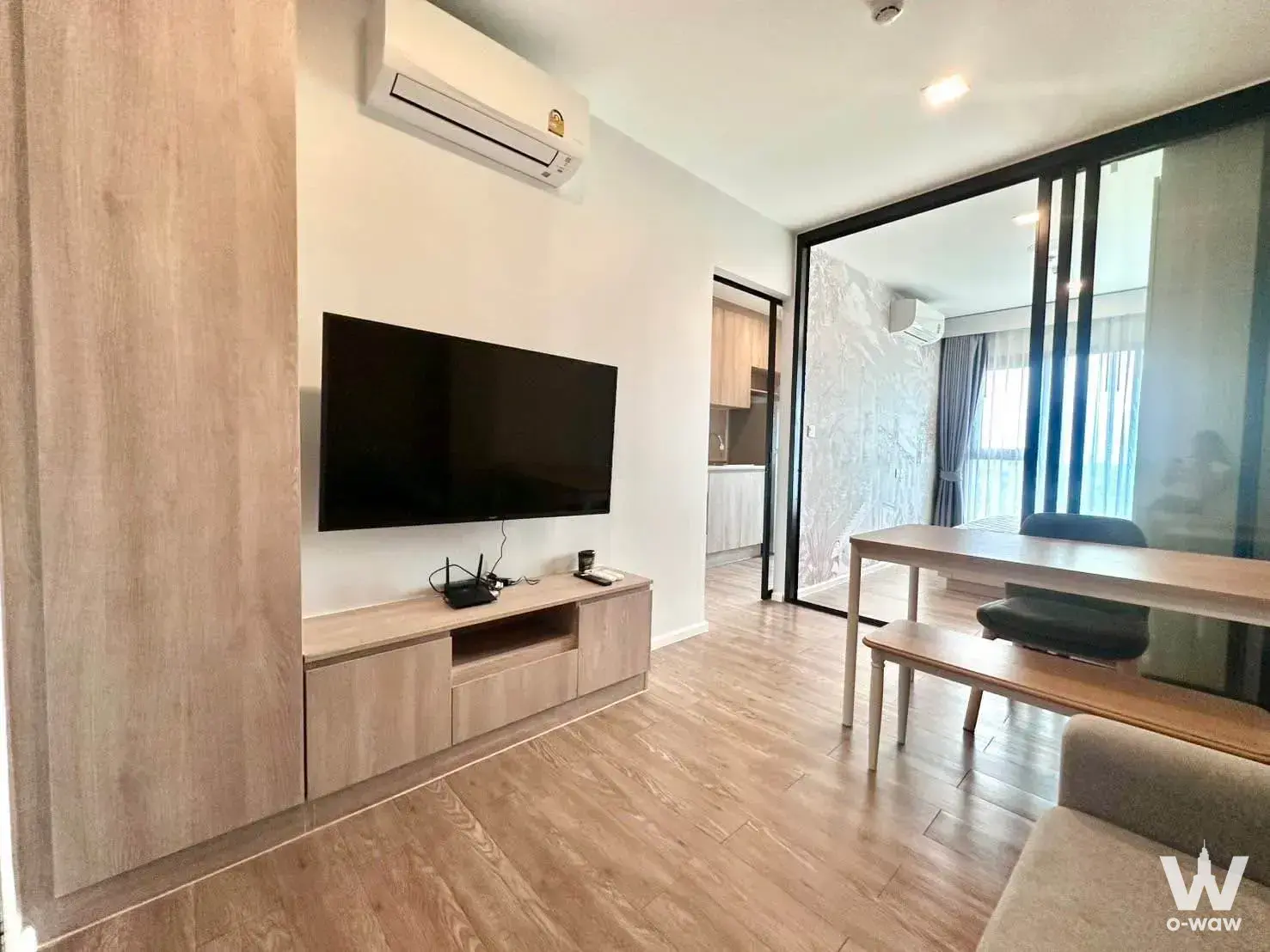 Living room in Modiz Sukhumvit 50 condo featuring wall-mounted TV, light wood paneling, and glass partition