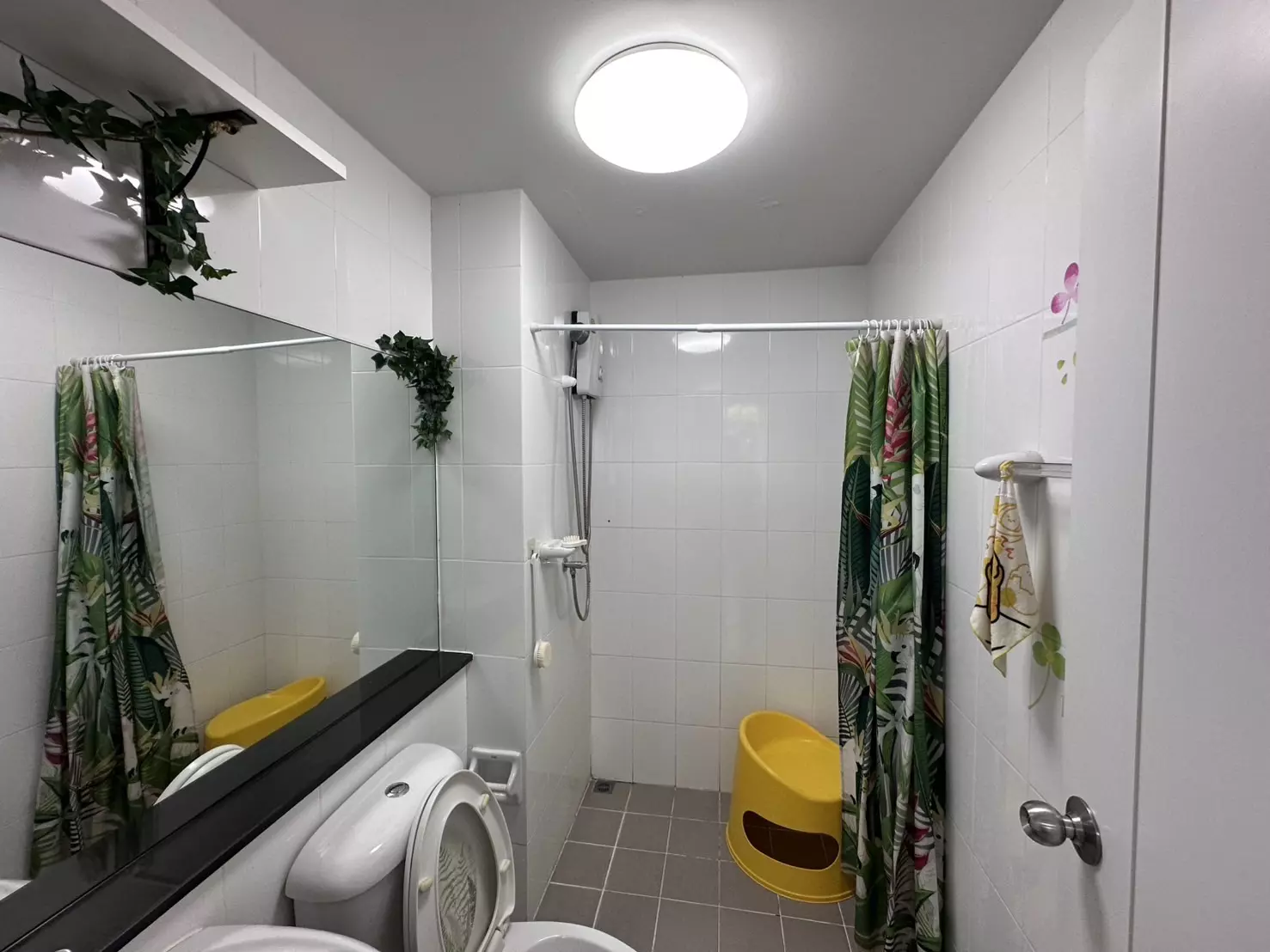 Compact PLUM Condo Phaholyothin 89 bathroom with white tiles, frameless mirror, and green tropical shower curtain