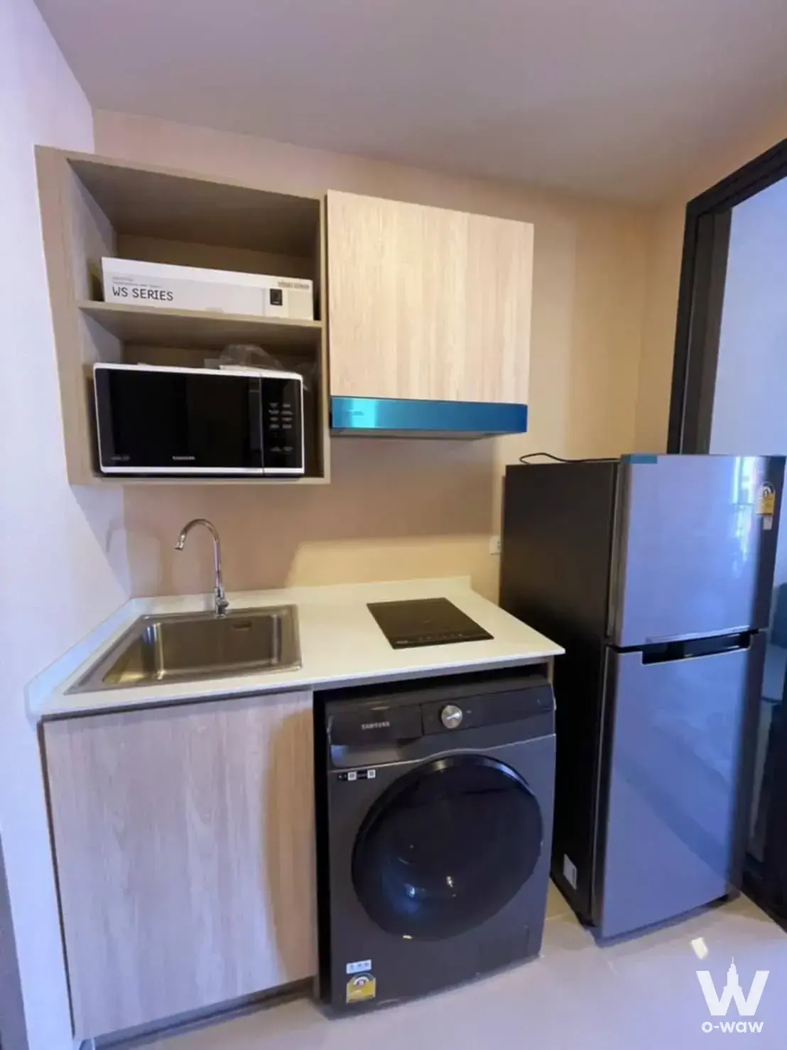 Compact kitchenette in The Muve Bangna featuring light wood cabinets and teal accent trim