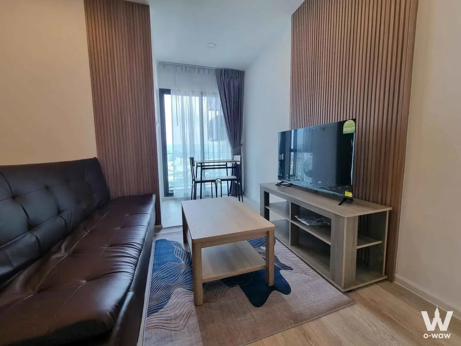 Modern living space in The Clev Riverline Chaophraya condo with dark leather sofa, wood slat accents, and city views