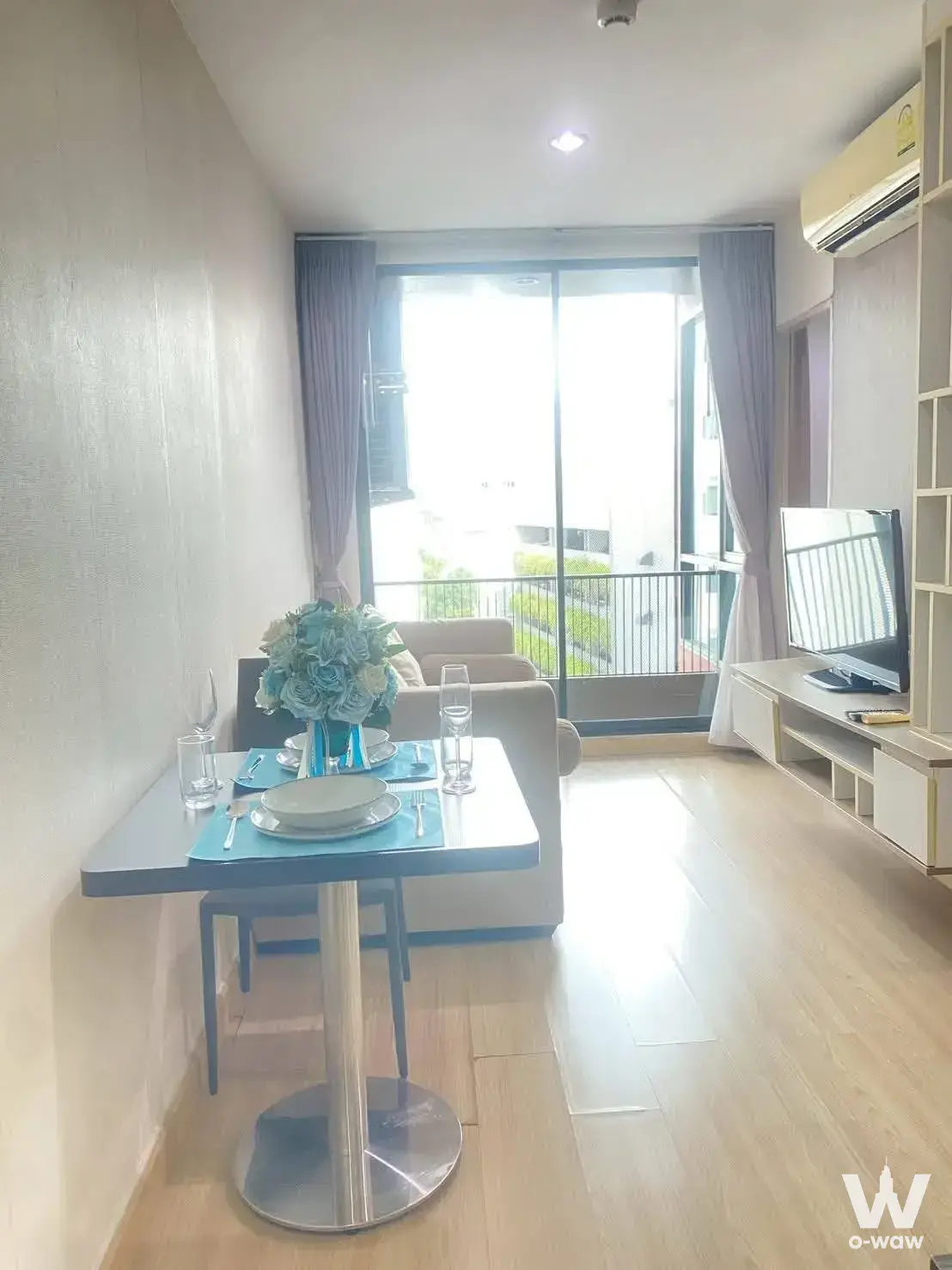 Living and dining room in The Unique Lat Phrao 26 condo featuring light wood flooring, teal accents, and balcony access